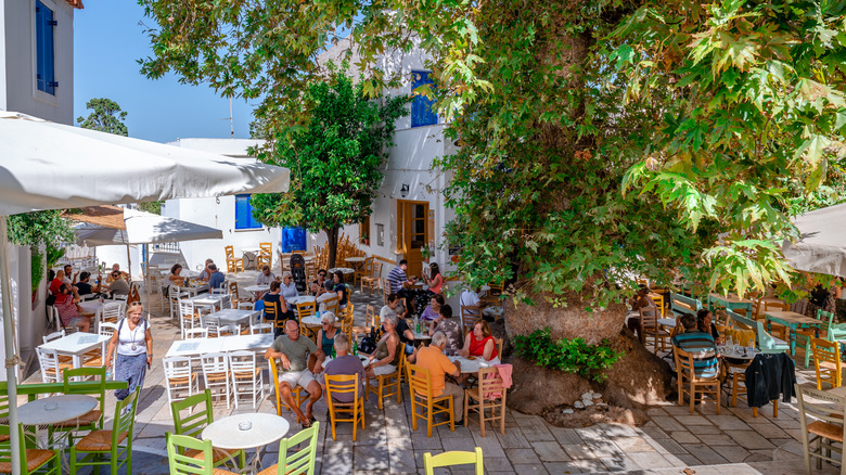 A busy main square with restaurant tables under a tree against whitewashed buildings with blue shutters