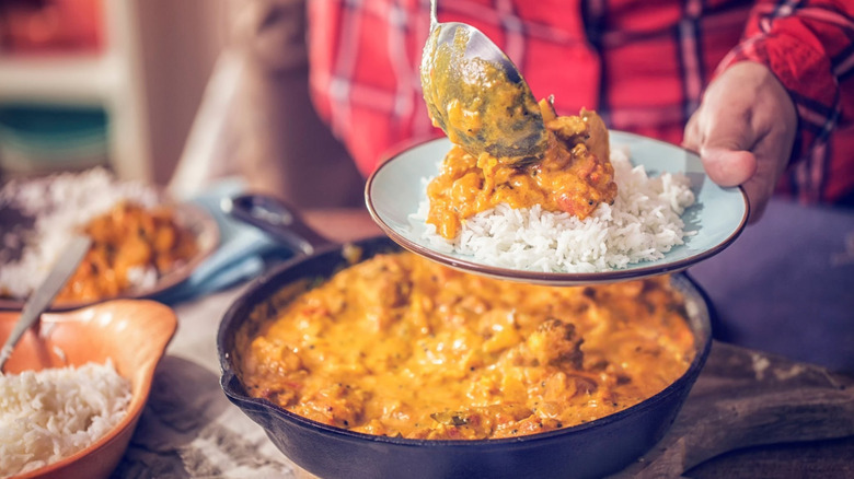 A man dressed in a red plaid shirt serving warm Indian-style curry over steaming rice on a plate with a large metal serving spoon