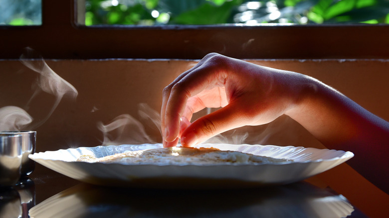 Close-up of a woman's right hand reaching for a dosa, an Indian flatbread typically eaten at breakfast, served on a porcelain plate, with light streaming through a window in the background