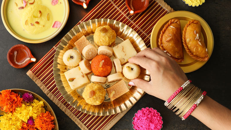 Close-up of a woman's hand and arm, adorned with elegant bangles, taking Diwali sweets off of a shared plate in India