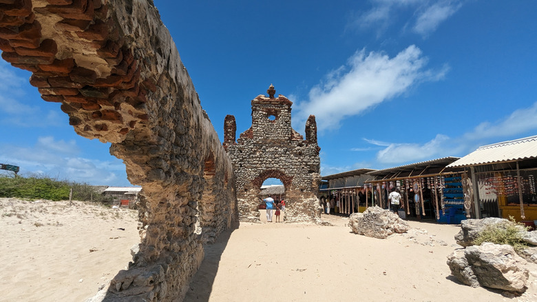 Facade and walls of an abandoned church on the sand against blue sky, with boulders and shacks next to it