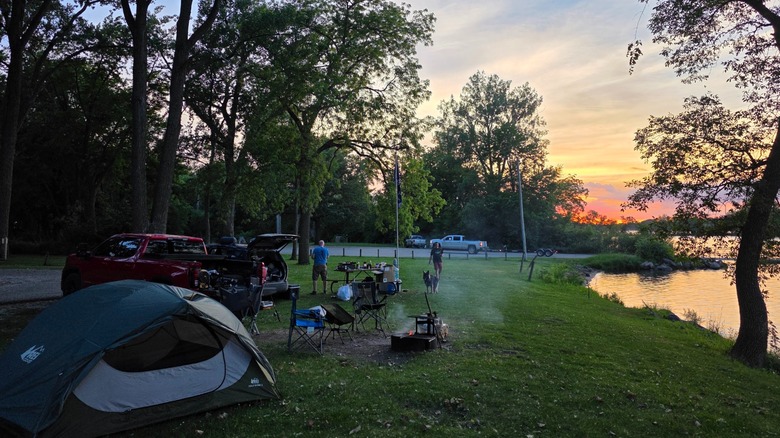 Waterfront campsite on a grassy area during sunset, with a park vehicle, picnic table, fire rings, and chairs at Black Hawk State Park, Iowa