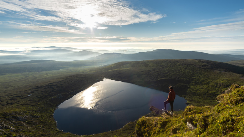 Man standing atop mountain overlooking a heart-shaped Lough Ouler lake in Wicklow, Ireland as the sun shines in the distance