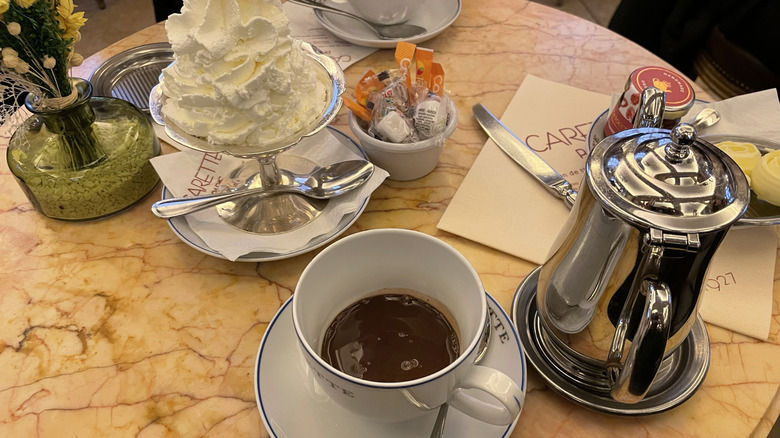 Cafe table setting with flowers, a cup of hot chocolate, a bowl of whipped cream, and a silver pot