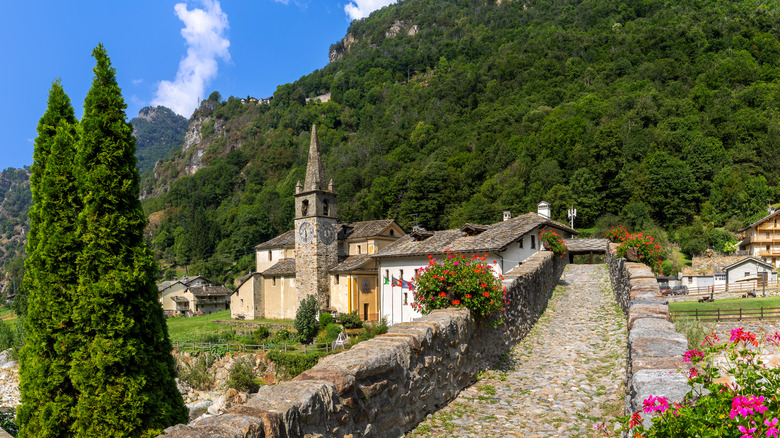 A picturesque view of a historic stone bridge leading to a church and houses surrounded by mountains and flowers in Aosta Valley, Italy