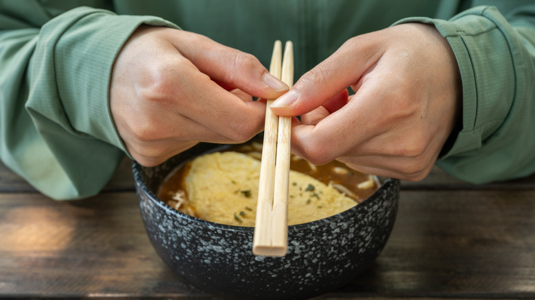 A close-up of a person in a green shirt holding chopsticks over a bowl of omelette