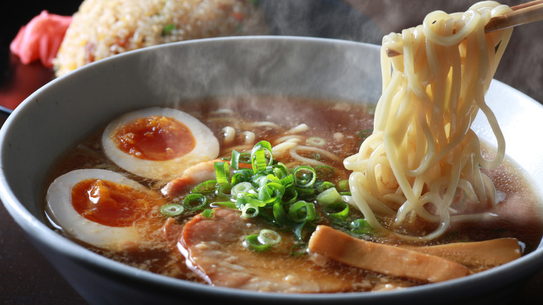 Bowl of steaming ramen with eggs, meat, and noodles, chopsticks going in for noodles and rice in the background