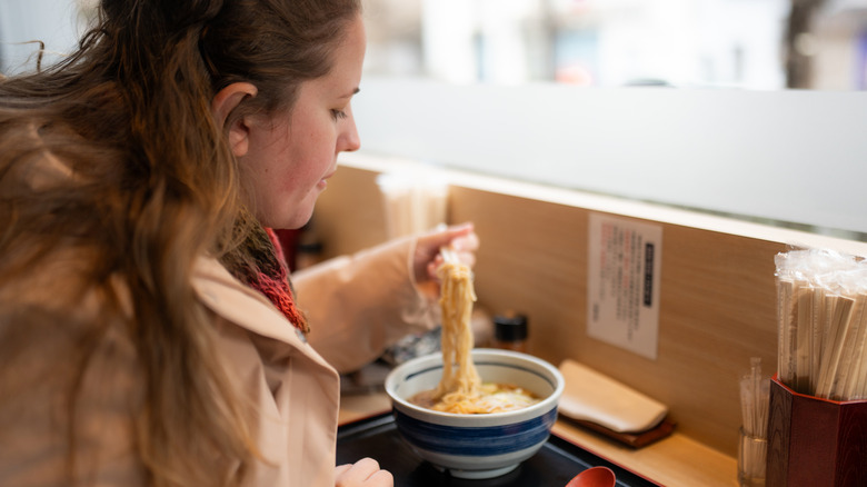 Woman dining on ramen at a restaurant counter in front of a window, noodles in chopsticks
