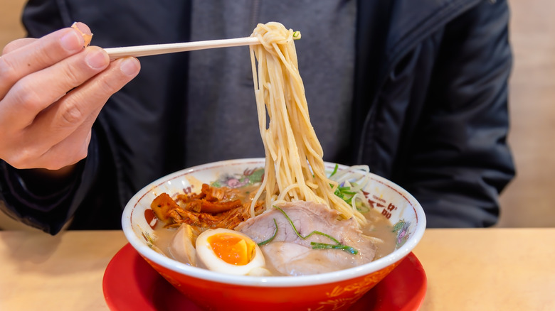 A red ramen bowl with egg and meat, plus long noodles being picked up with chopsticks