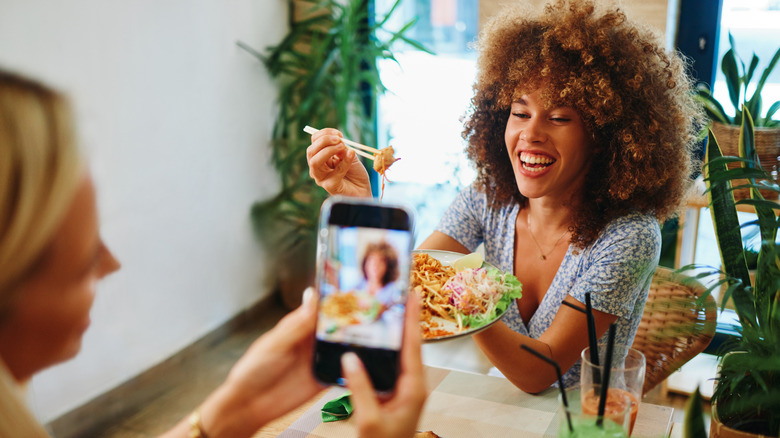 Two friends at lunch, one taking a photo of the other eating with a cell phone