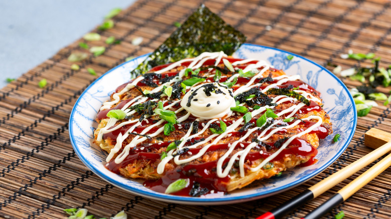 A plate of okonomiyaki, a Japanese noodle pancake, drizzled in mayonnaise and brown sauce and scattered with chopped green onions