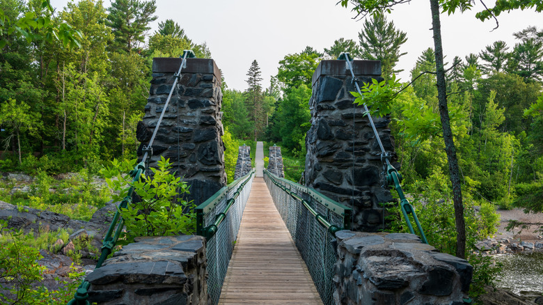 A photo from the start of the pedestrian suspension bridge with stone pillars and green trees all around