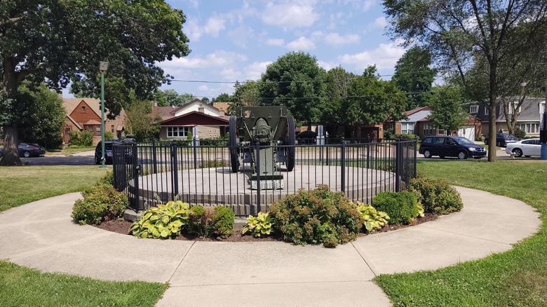View from Memorial Park in Blue Island, with a fenced-in canon in the foreground and suburban homes across the street, surrounded by trees under a cloudy blue sky