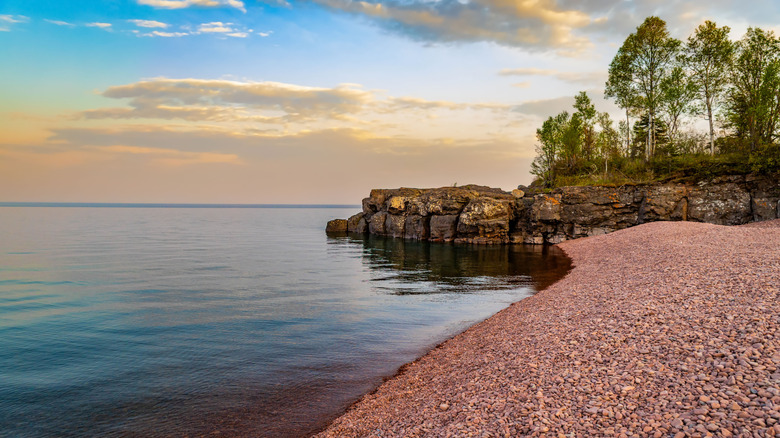 A photo on Iona's Beach showing the pink stones, rugged rocks on the end of the beach topped with trees, and evening light on the calm water