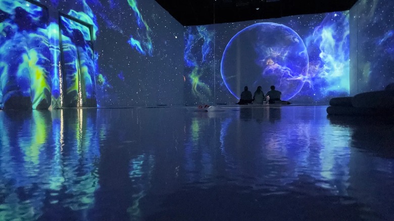 A room with projections on the wall inside the museum and three people sitting on the floor looking at it