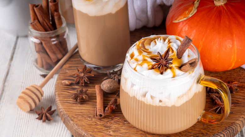 A pumpkin spice latte in a glass mug on a wooden tray with a pumpkin, cinnamon sticks, and more autumn decor around it