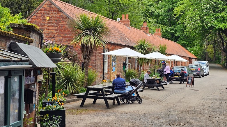 Picnic tables next to a red brick one-story buildings with people enjoying a meal and greenery all around