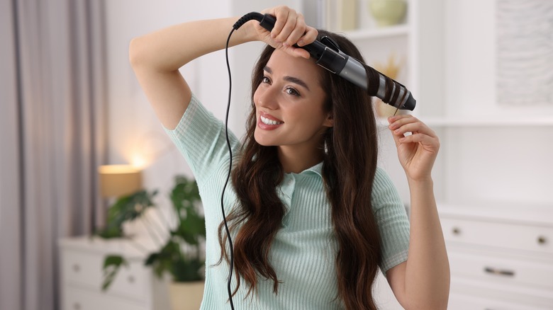 a woman smiling as she uses a curling iron to style her hair