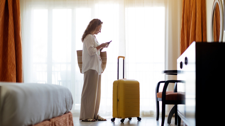 a woman looks into her phone in front of a brightly lit window, with a yellow suitcase on the floor in front of her