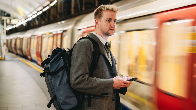 A person in business clothing wearing a backpack while holding a phone as a train comes into the London Underground station