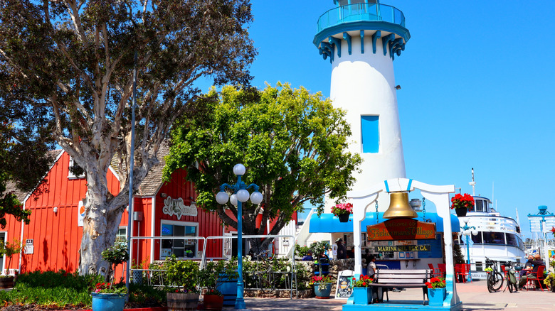 Red building, blue and white lighthouse, white bench, and gold bell flanked by trees at Fisherman's Village in Marina del Rey