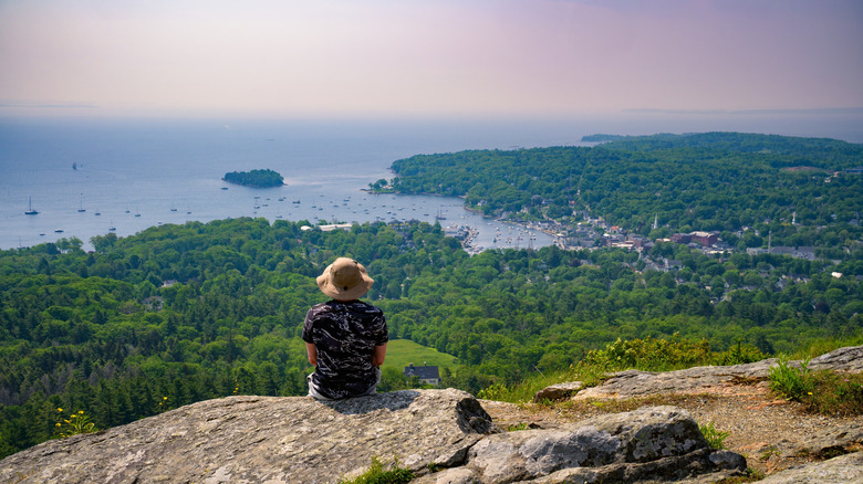 A hiker sits on a rocky ledge at the summit of Mt. Battie in Camden Hills State Park, overlooking Camden Harbor and Penobscot Bay in Maine