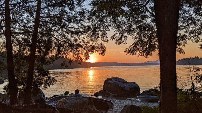 Sunset over a the calm Sebec Lake framed by tall trees, with kayaks and large rocks along the sandy shoreline.