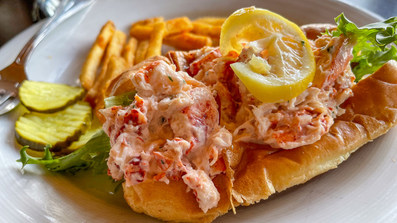 A lobster roll with a lemon slice next to french fries and pickles on a plate
