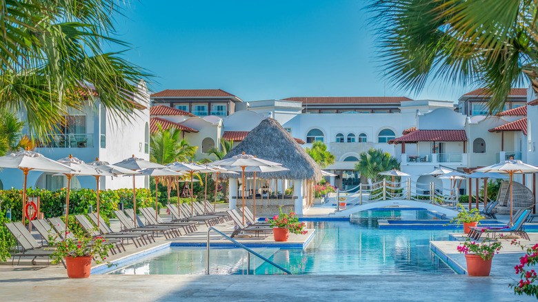 View of a beautiful resort in the Caribbean on a clear blue summer day, with a calm pool, lounge chairs, umbrellas, flower pots, and palm trees visible.
