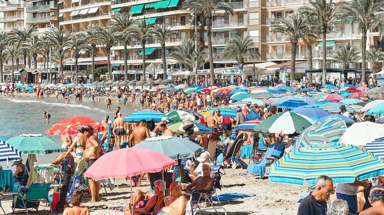 People sunbathe on a beach in Playa del Cura in Alicante, Costa Blanca, Spain, with summer apartments in the background. It is a warm, sunny day, and the beach is packed with people and colorful umbrellas.