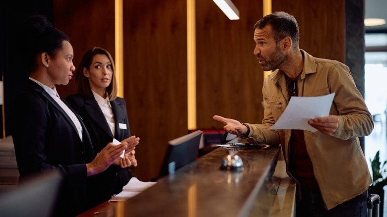 An angry guest discusses his bill with two female staff working at the concierge of a modern hotel.