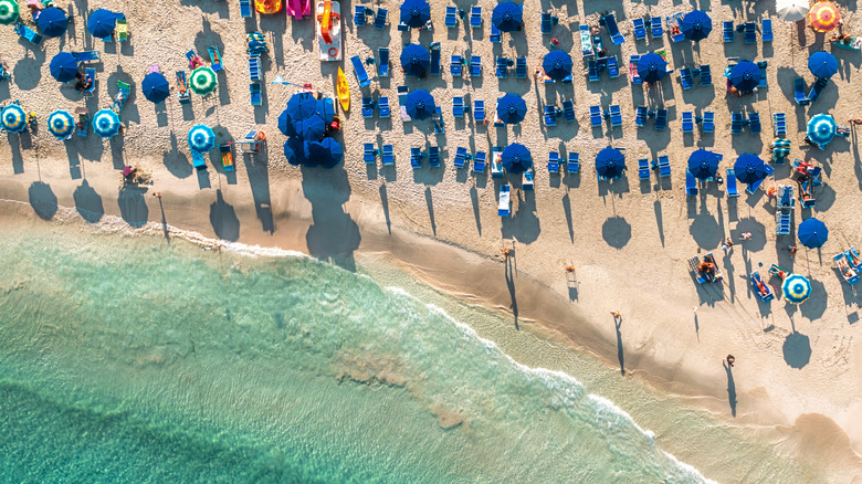 Aerial view of a beach covered in blue umbrellas and many people, with turquoise waters lapping at its shores, on a hot, bright day.