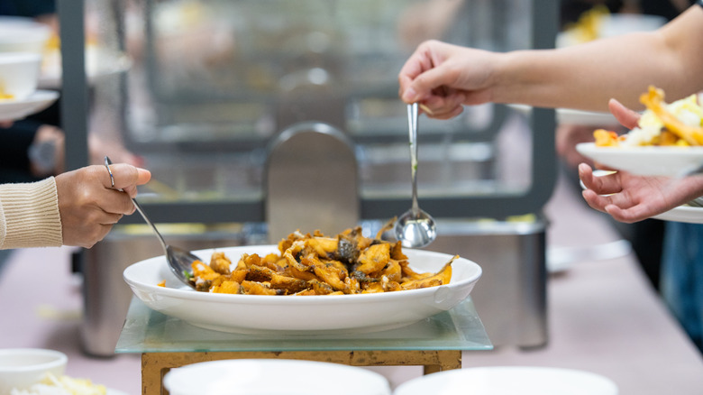 Close-up of two people using spoons to grab food off of a plate on a buffet table at a hotel.