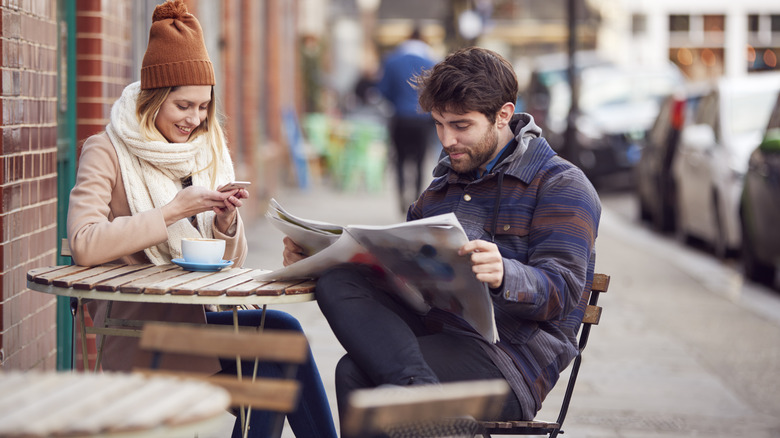Couple reading newspaper and checking phone at a cafe table