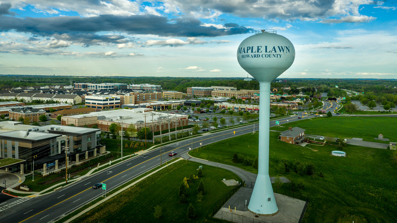 Blue water tower reading Maple Lawn Howard COunty, surrounded by green grass with commercial buildings and homes on the left side of the frame, under a cloudy blue sky