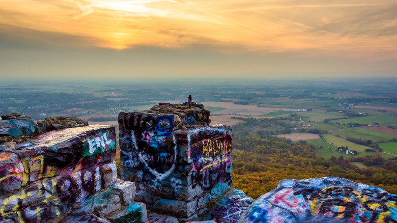 A hiker perched atop High Rock, a graffiti-covered scenic overlook in Maryland, featuring a vast valley and a golden sunset..