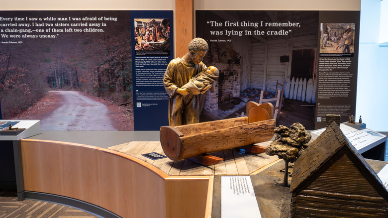 Exhibits inside the Visitor Center, with a bronze statue of a man holding a baby in the center behind a wooden crib, with a small model house in teh foreground and signs with words behind it
