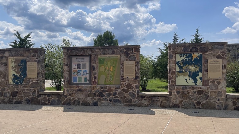 Stone walls with signs outside the Visitor Center, with trees and grass in the background, under a cloudy blue sky