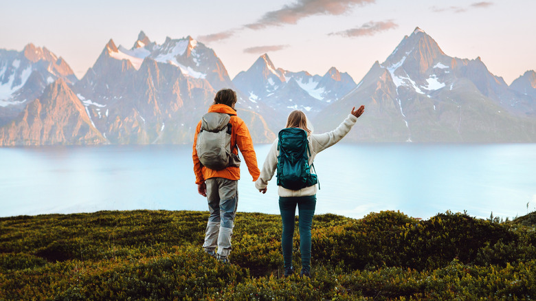 A couple holding hands and hiking in the Lyngen Alps in Norway, their backs toward the camera, with a lake and a mountain range visible in the background.