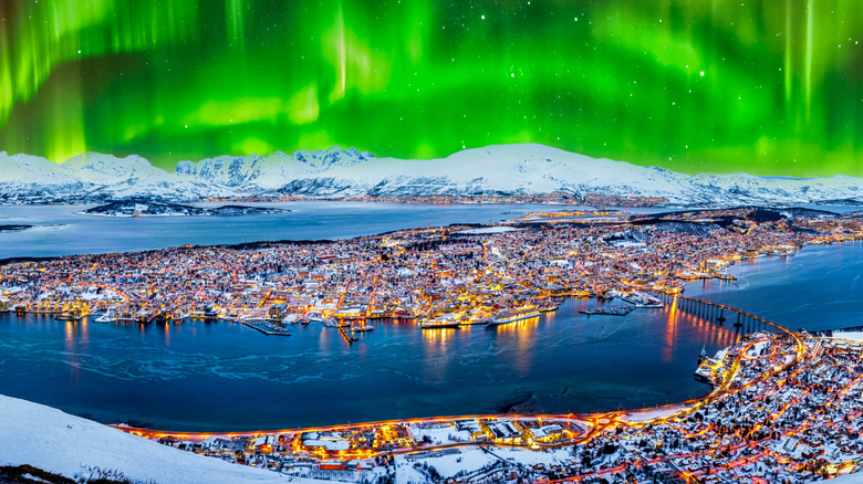 A panoramic view of the city of Tromso, Norway, with snow-covered mountains in the background and the sky illuminated by the twinkling Northern Lights.