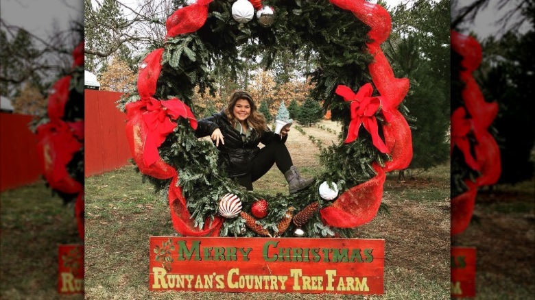 Woman posing in front of a large decorated wreath with a sign at Runyan's Country Tree Farm