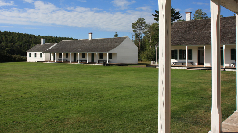 Two large one-story white wooden buildings from inside Fort Wilkins with green grass in front and a hill with pine trees behind