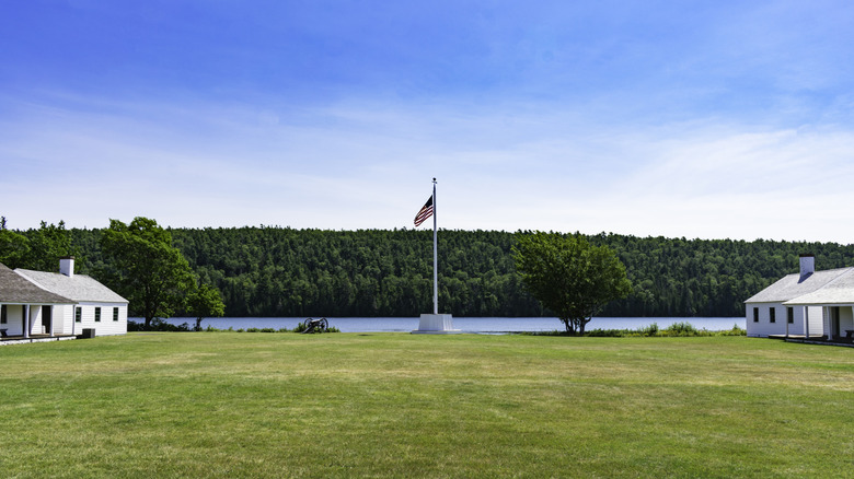 An American flag on a pole at the center of the grounds at Fort Wilkins, with old buildings on either side and a calm, tree-lined lake behind it