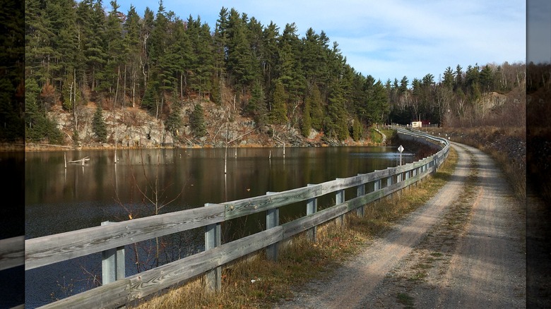 The Iron Ore Heritage Trail running along a small calm lake with a wooden fence and a rocky pine-covered hill on the far shore
