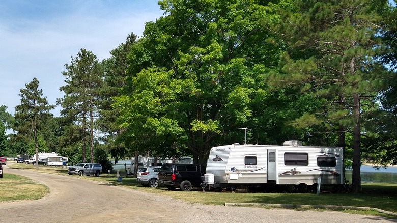 A road at the campground in Metamora-Hadley, with a few campers, vehicles, tall trees, and the lake behind them to the right
