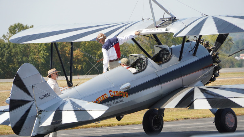 Close up of a small biplane from the Bealeton Flying Circus Air Show with two people on the plane and one standing on the ground behind.