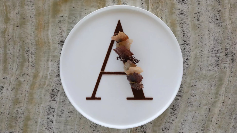 Chocolate dessert in the shape of an "A" with edible decorations on one side of the letter, all on a white plate and marble table.