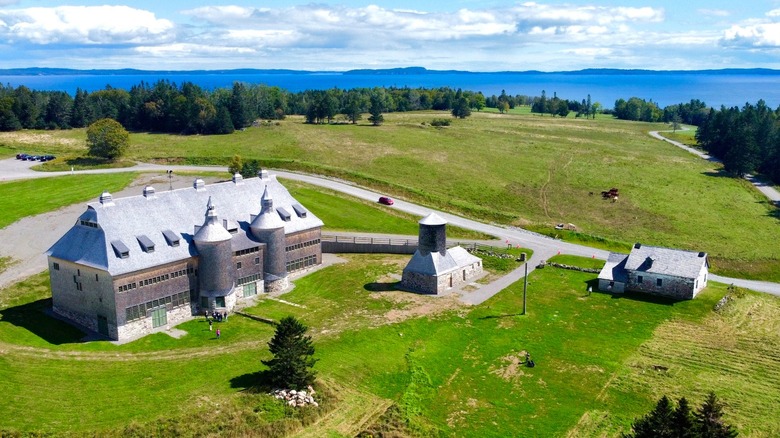 a historic barn on green fields with pine trees and coastline in the background