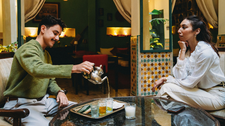 A man pouring Moroccan mint tea from a silver tea pot seated across a table from a woman.