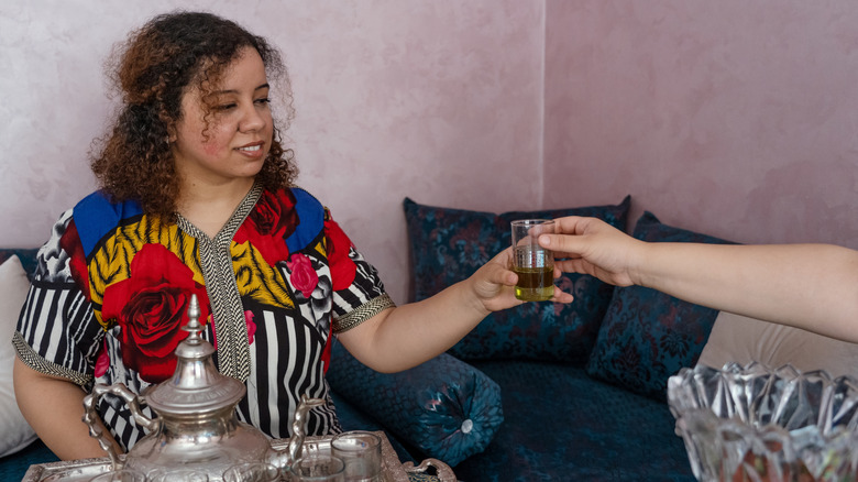 A hand passing a glass of mint tea to a woman sitting on a couch dressed in a colorful outfit